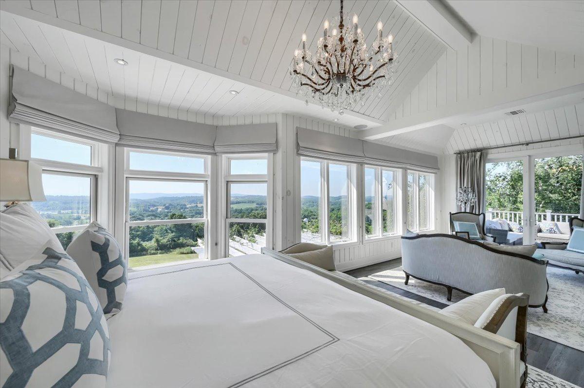 Primary bedroom at The Maple Overlook — king bed, crystal chandelier, shiplap ceiling, panoramic Hudson Valley views through floor-to-ceiling windows, private balcony